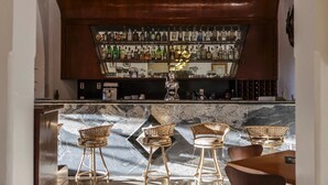 Un espace bar doté d’un comptoir en marbre, de tabourets de bar en osier et de différentes bouteilles d’alcool exposées sur des étagères.