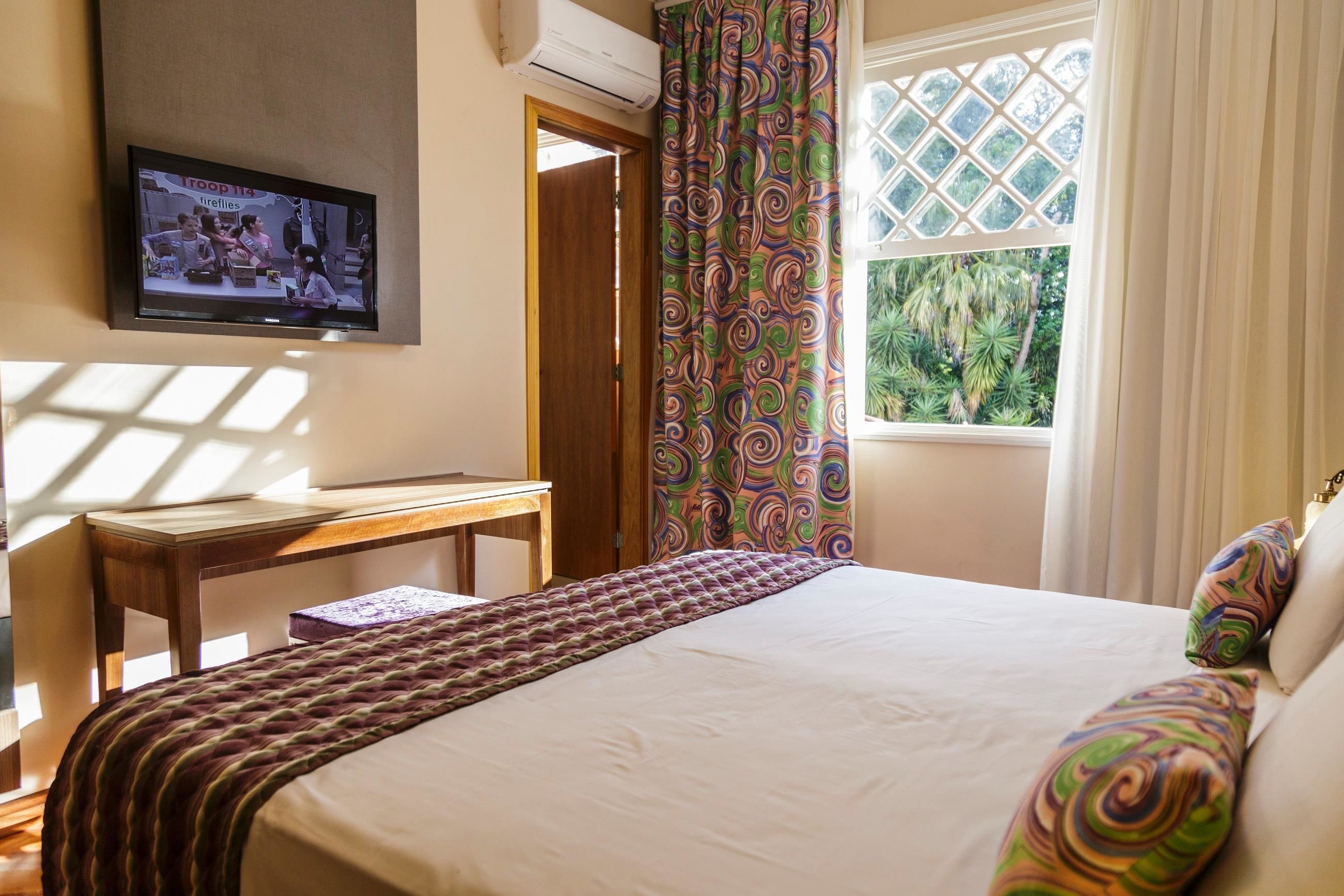 Una camera d'albergo con un letto, una TV fissata al muro, una finestra con tende e una scrivania con una sedia.