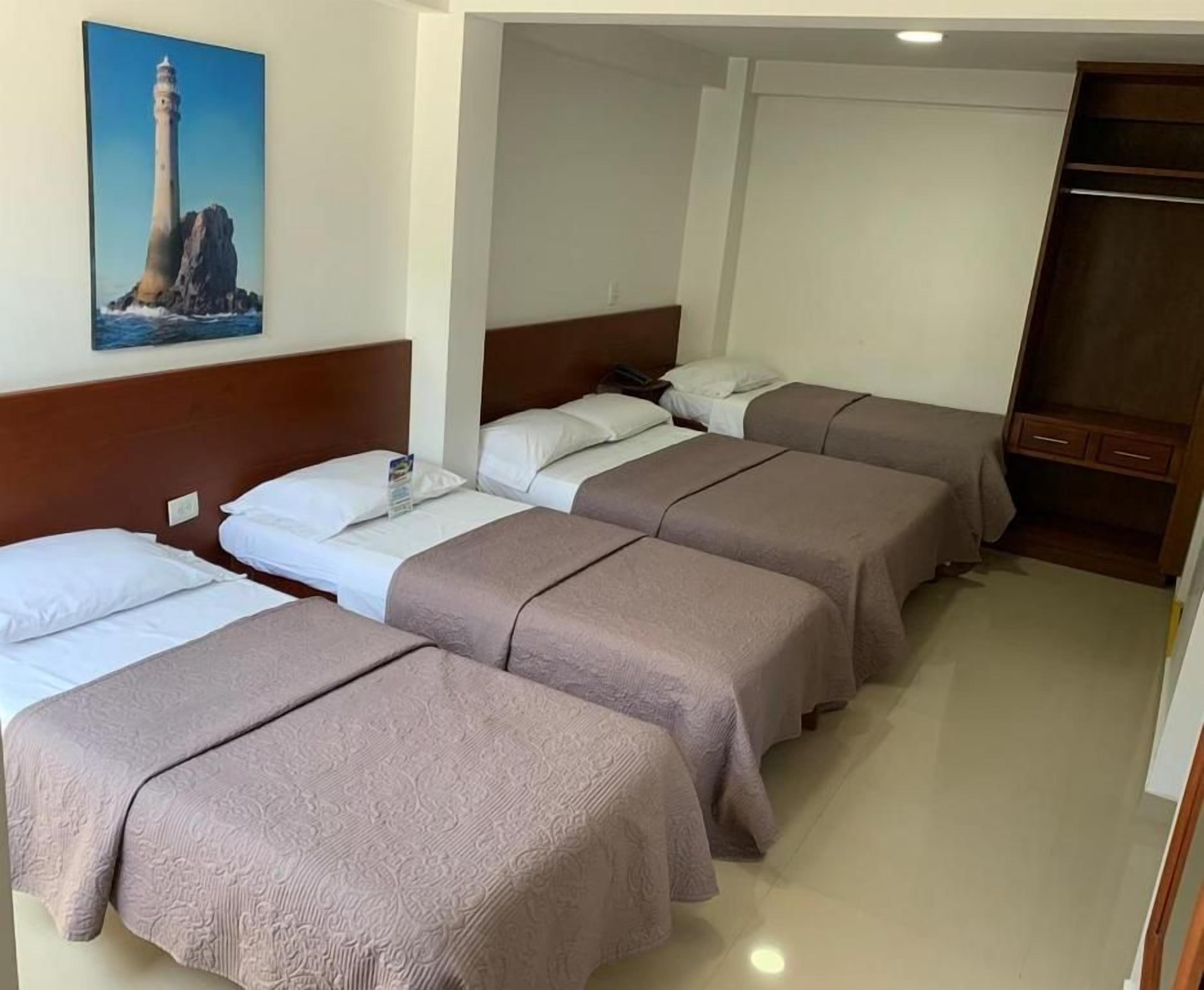 Une chambre d’hôtel avec trois lits, une tête de lit en bois, un tableau représentant un phare et une armoire.