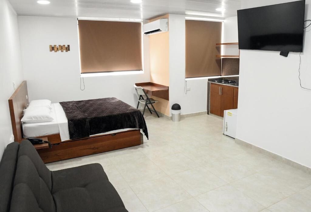 Una habitación pequeña con cama, escritorio, silla, televisor y kitchenette.