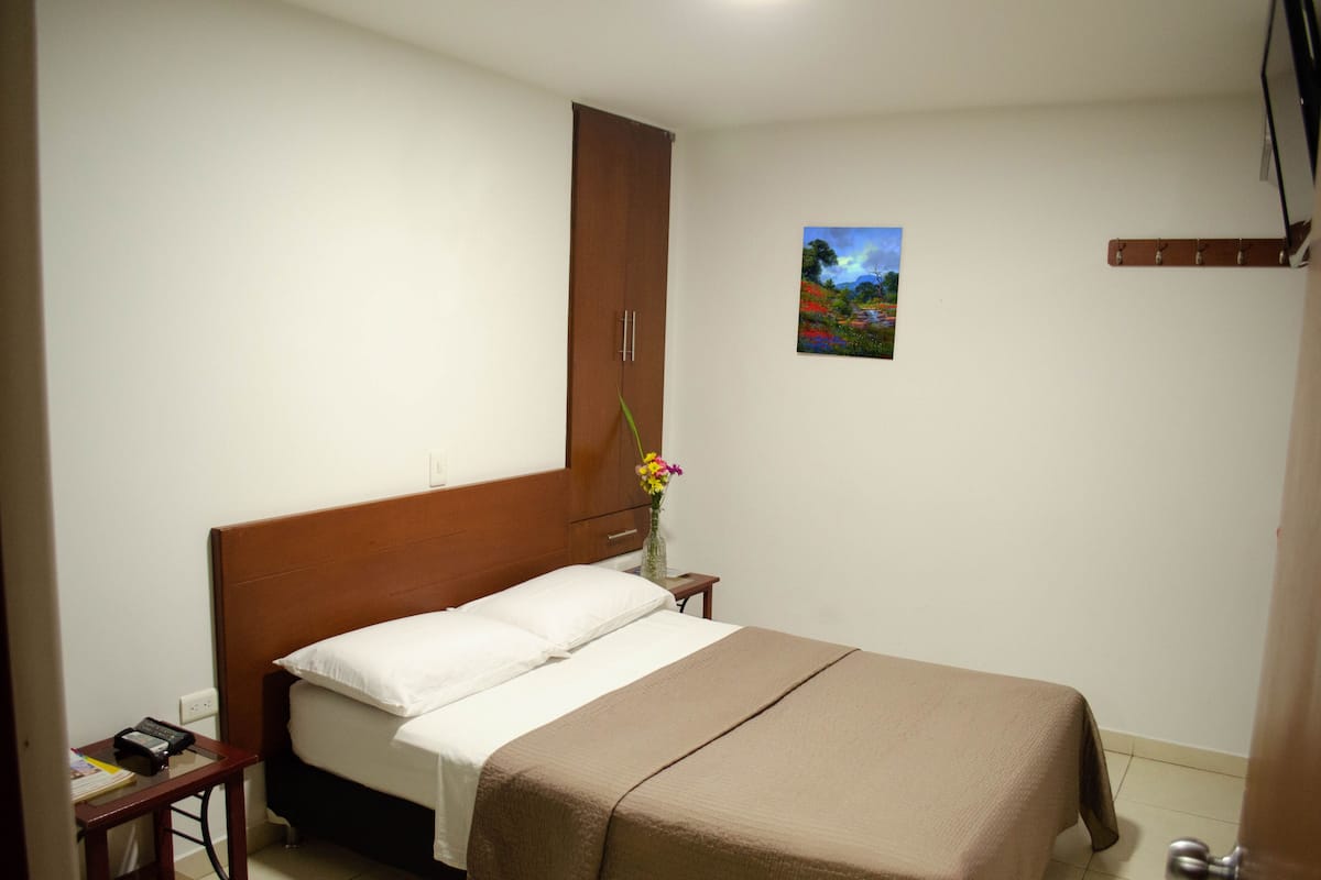 A hotel room with a bed, a nightstand, a wooden wardrobe, a small table with a vase of flowers, and a painting on the wall.
