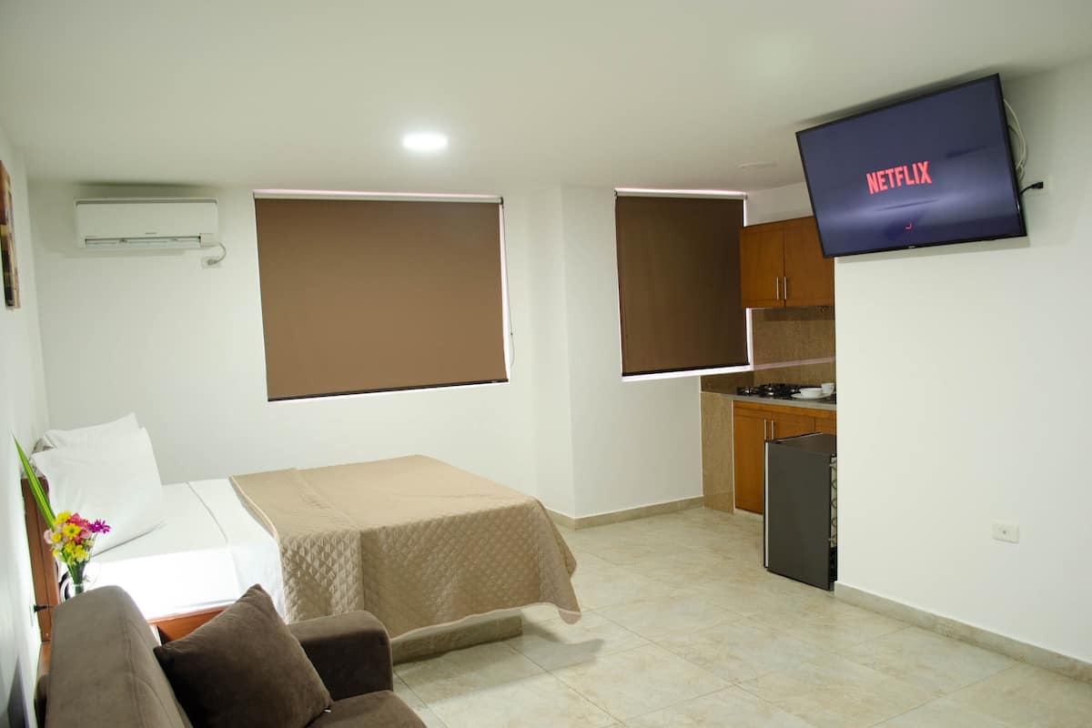 A hotel room with a bed, a sofa, a TV mounted on the wall, a kitchenette, and a window with blinds.