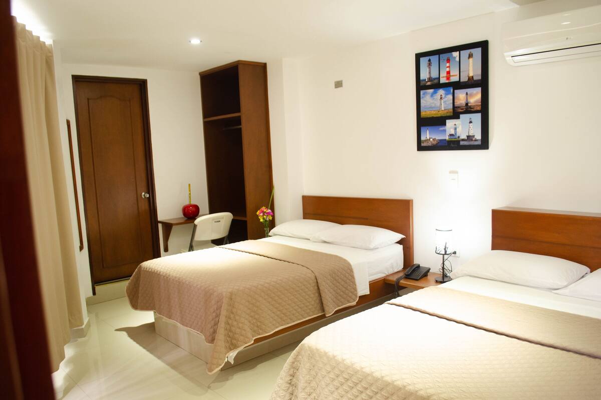 A hotel room with two beds, a wooden wardrobe, a desk with a lamp, and a wall-mounted photo collage.