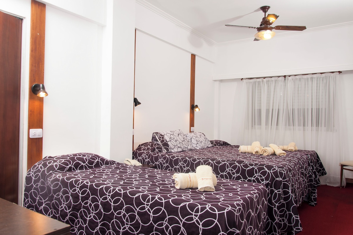 A hotel room with two beds, a ceiling fan, a window with sheer curtains, and a wooden door.