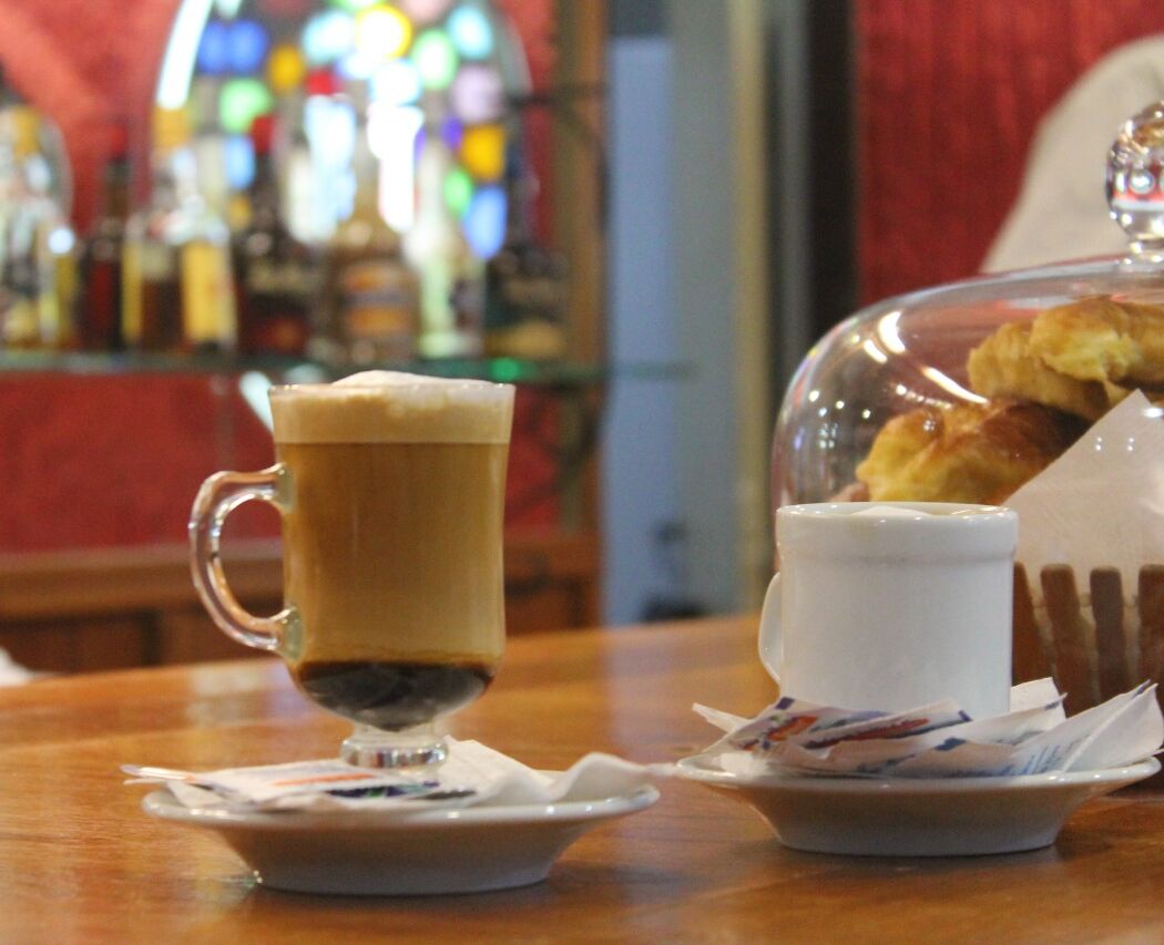 A coffee cup on a saucer, a small plate with a pastry, and a glass dome covering a pastry display.