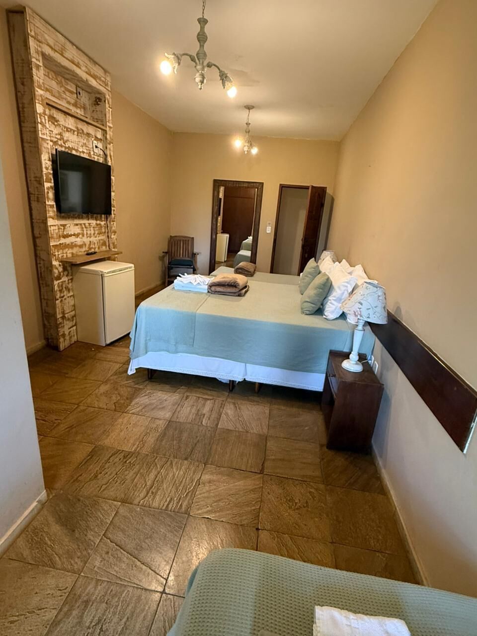 A hotel room with a bed, a small refrigerator, a television mounted on a wooden wall, a chandelier, and a mirror.
