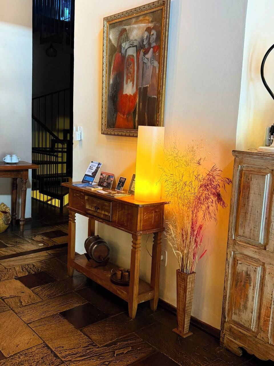 Ein Zimmer mit Holztisch, Stehlampe, gerahmtem Gemälde und dekorativer Vase mit Trockenpflanzen.