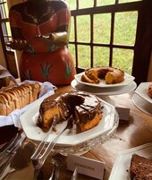 Uma mesa de sobremesas com diversos bolos e doces, um bule de chá e vista para a janela.