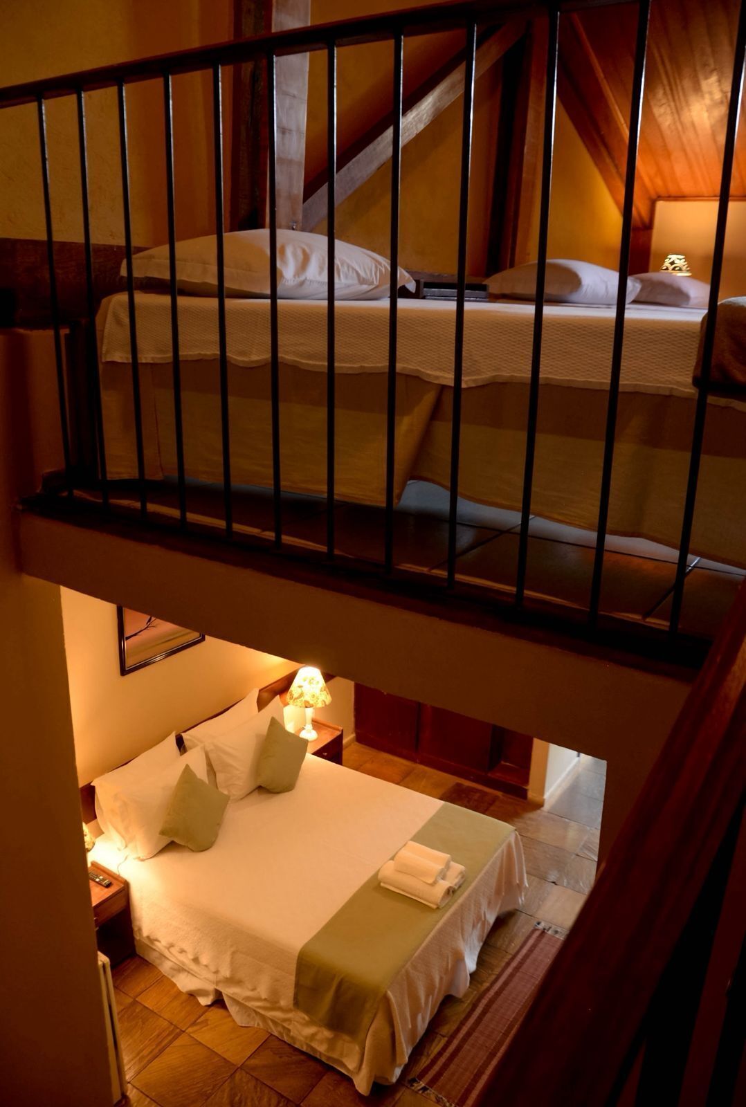 A two-level bedroom with a balcony, a bed with white bedding, a nightstand with a lamp, and a wooden floor.