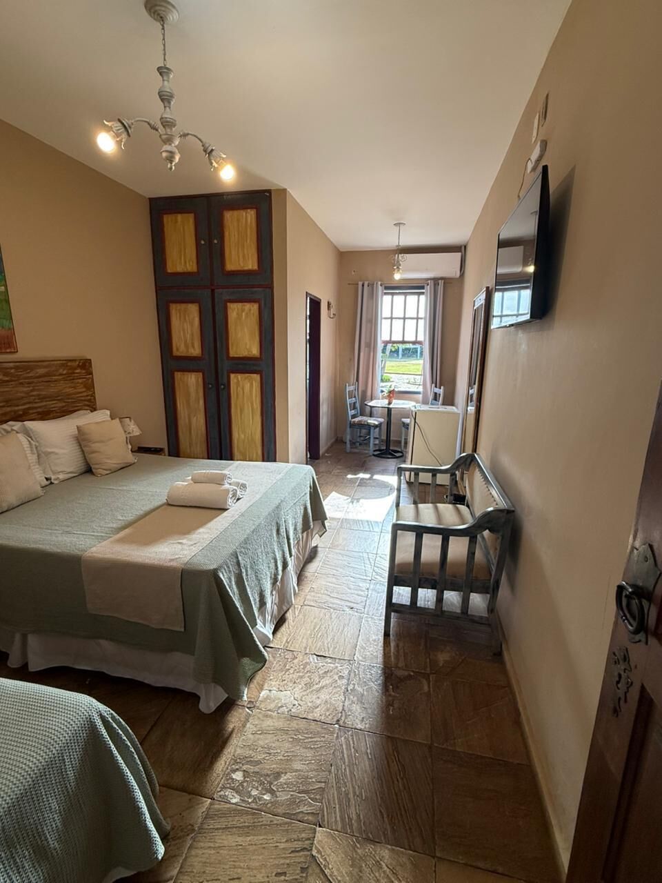 Une chambre d’hôtel avec un lit, une chaise, une table, un téléviseur et une fenêtre avec des rideaux.