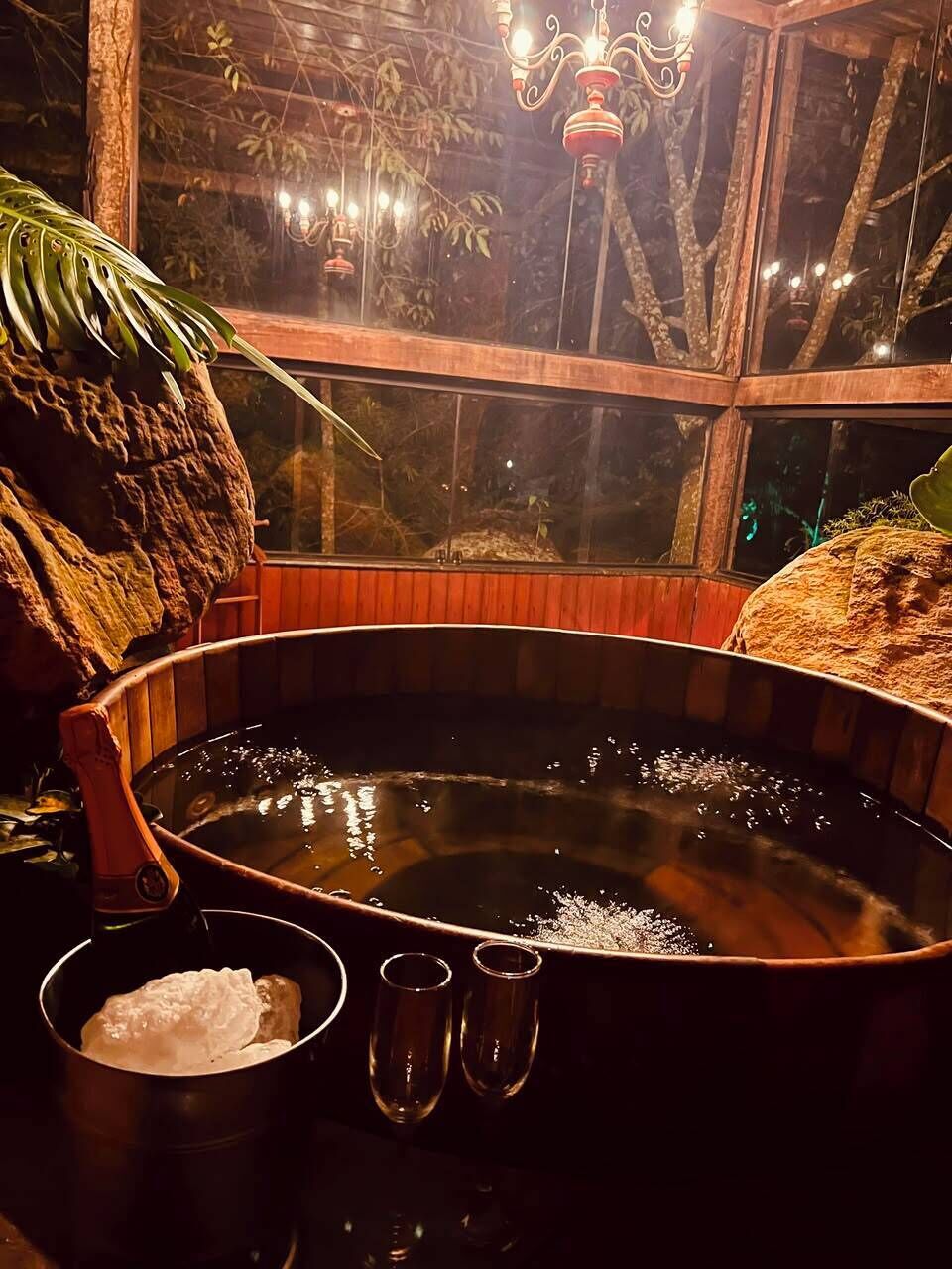 Uma banheira de hidromassagem com estrutura de madeira, uma janela com vista para árvores e um candelabro.