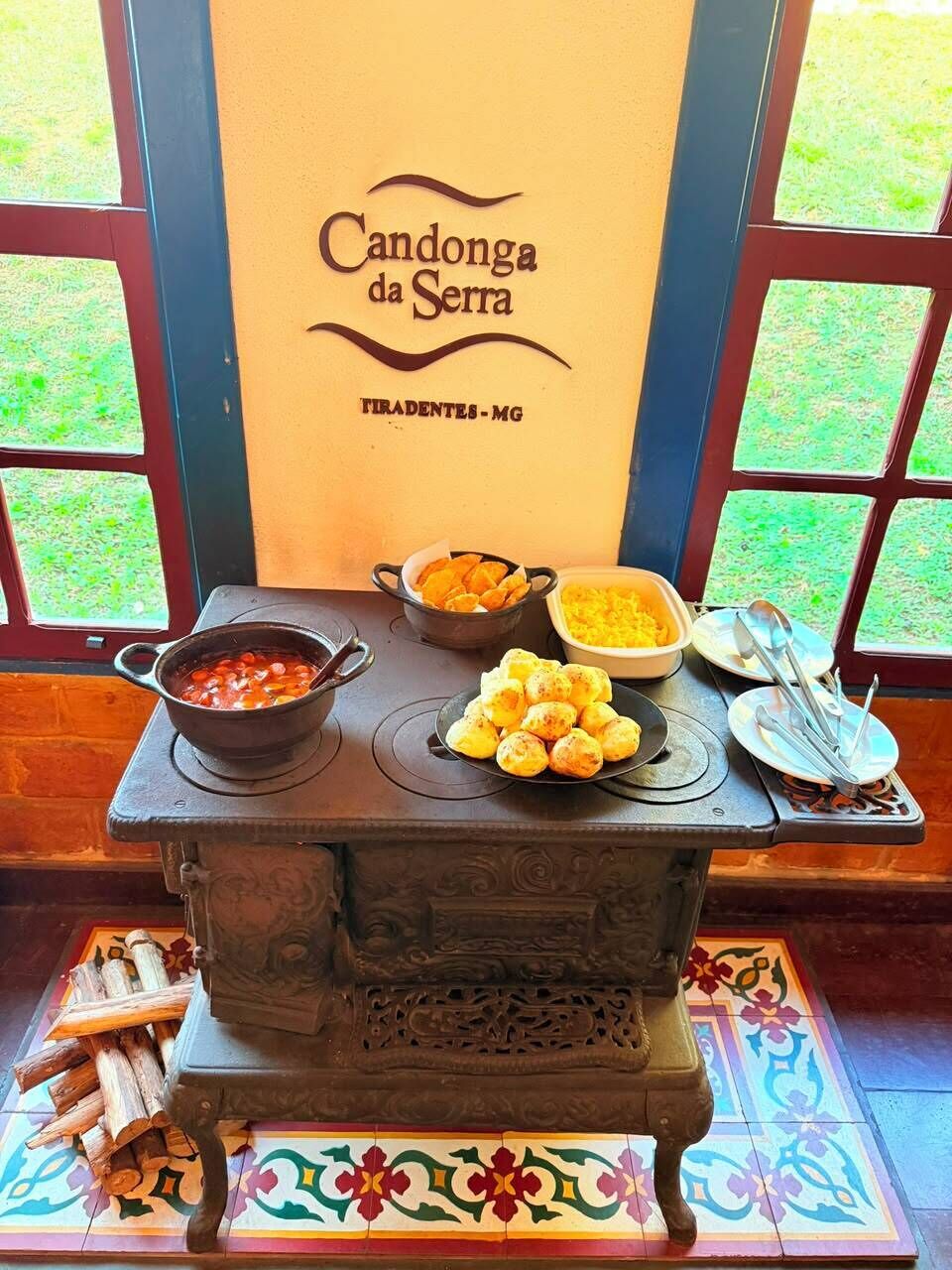 Estufa tradicional con una olla de comida, un tazón de frijoles y un plato de comida sobre una mesa con piso de baldosas y una ventana que da a la vegetación.
