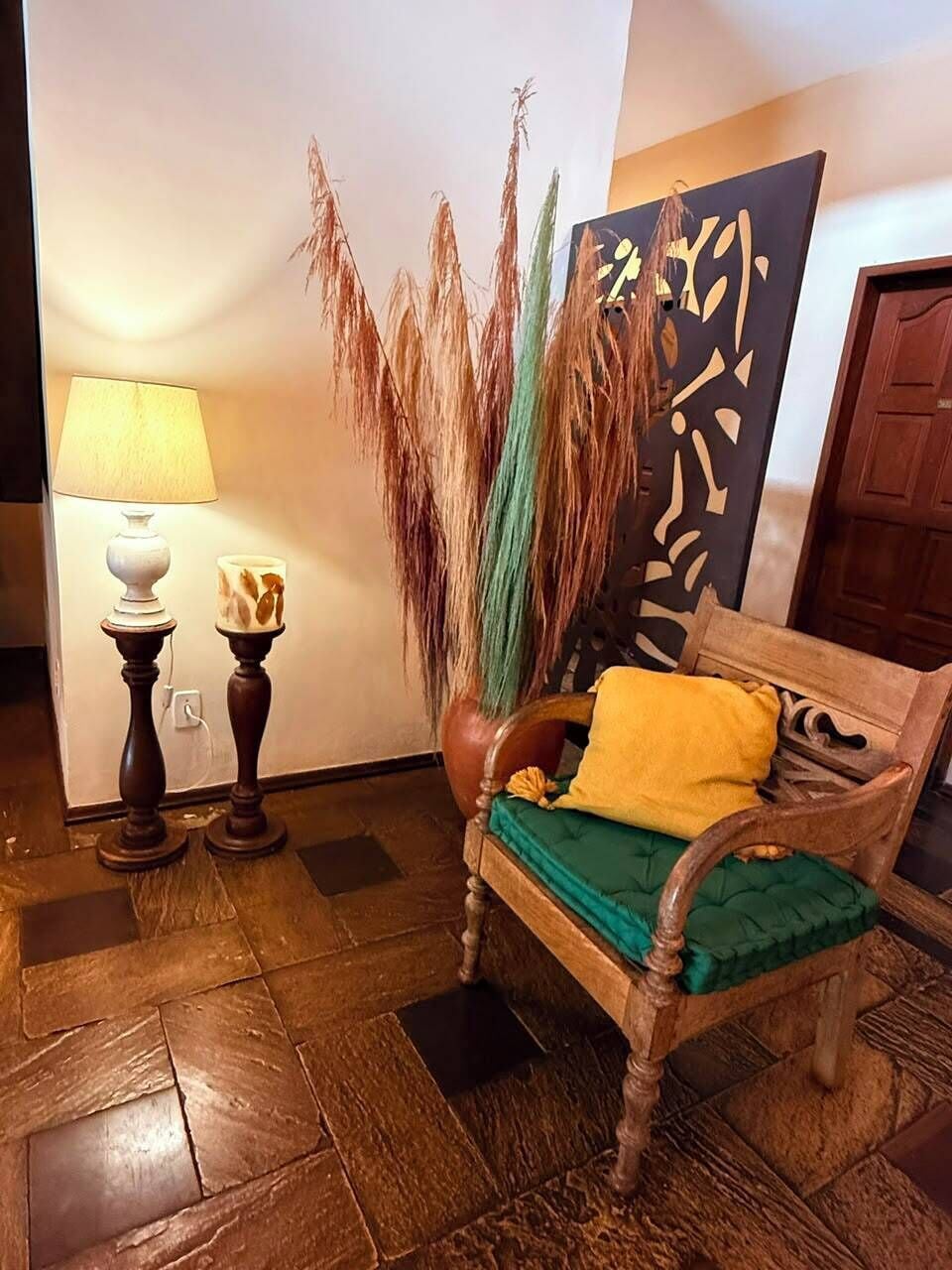 A wooden bench with a green cushion and a yellow pillow, a tall decorative plant, a wall-mounted abstract art piece, and two floor lamps with patterned lampshades.