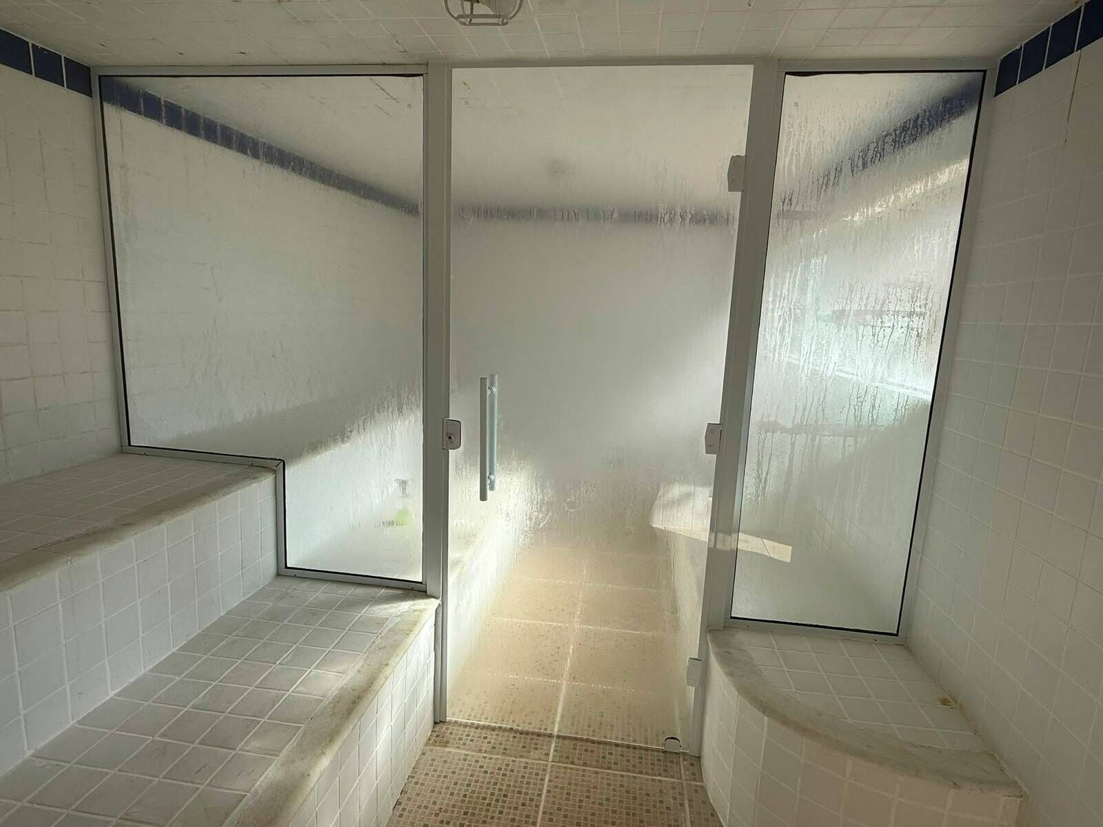 Une salle de bain avec des murs et un sol carrelés de carreaux blancs, une porte en verre dépoli et un petit banc.