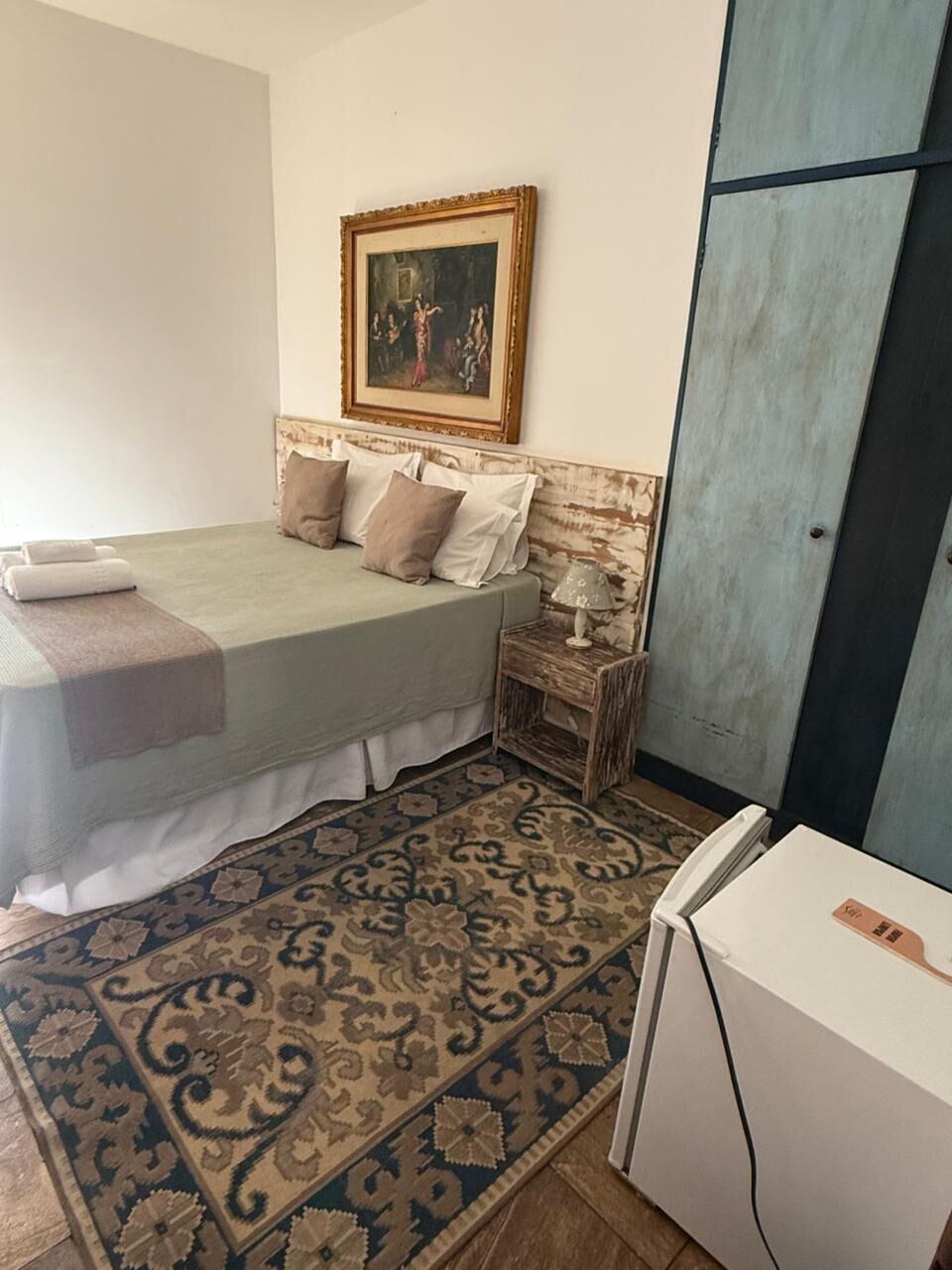 A bedroom with a bed, pillows, a nightstand with a lamp, a patterned rug, and a wardrobe.