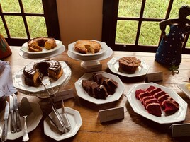 A dessert table with various cakes and pastries labeled with names like "BANANA" and "CENOURA".