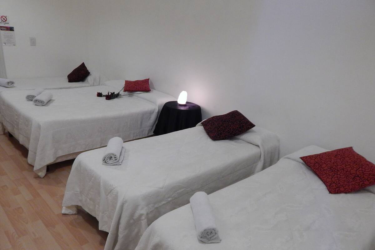Une chambre avec trois lits, chacun recouvert d’une courtepointe blanche et agrémenté d’une serviette pliée. Une petite table avec une lampe allumée se trouve entre les lits.