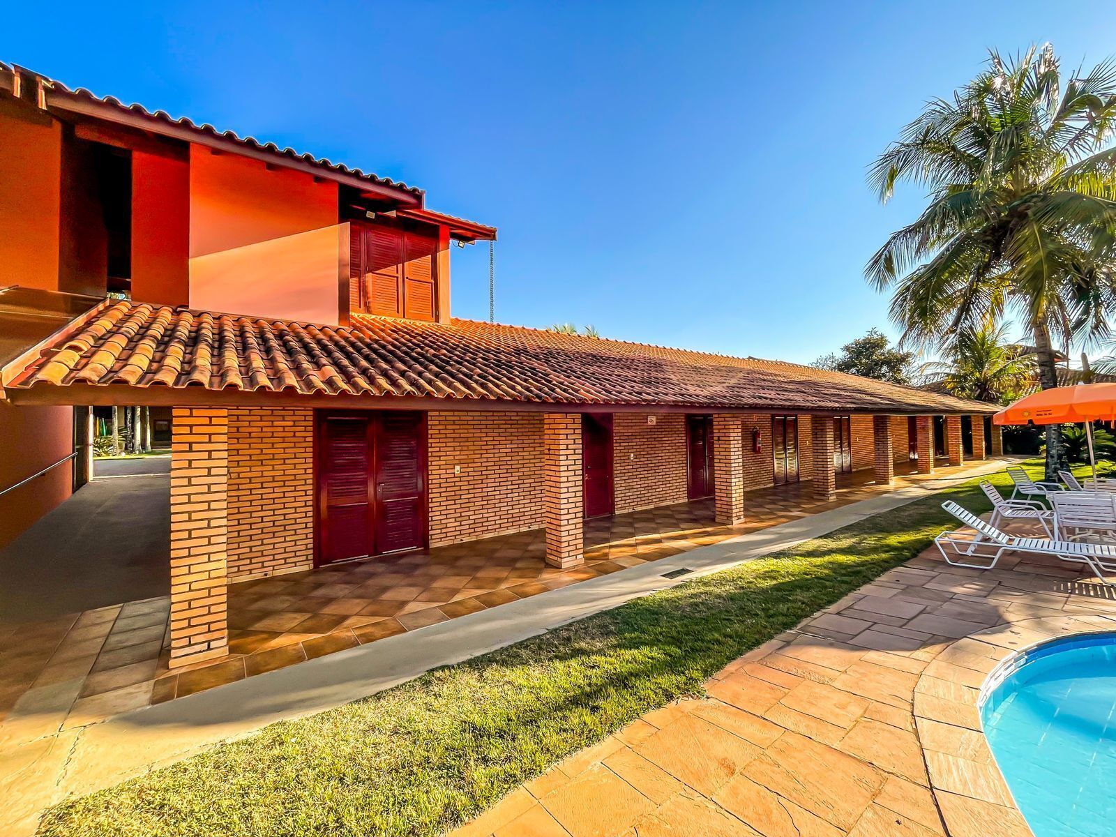 A two-story building with a tiled roof and brick walls, a swimming pool, and a covered patio area with lounge chairs.