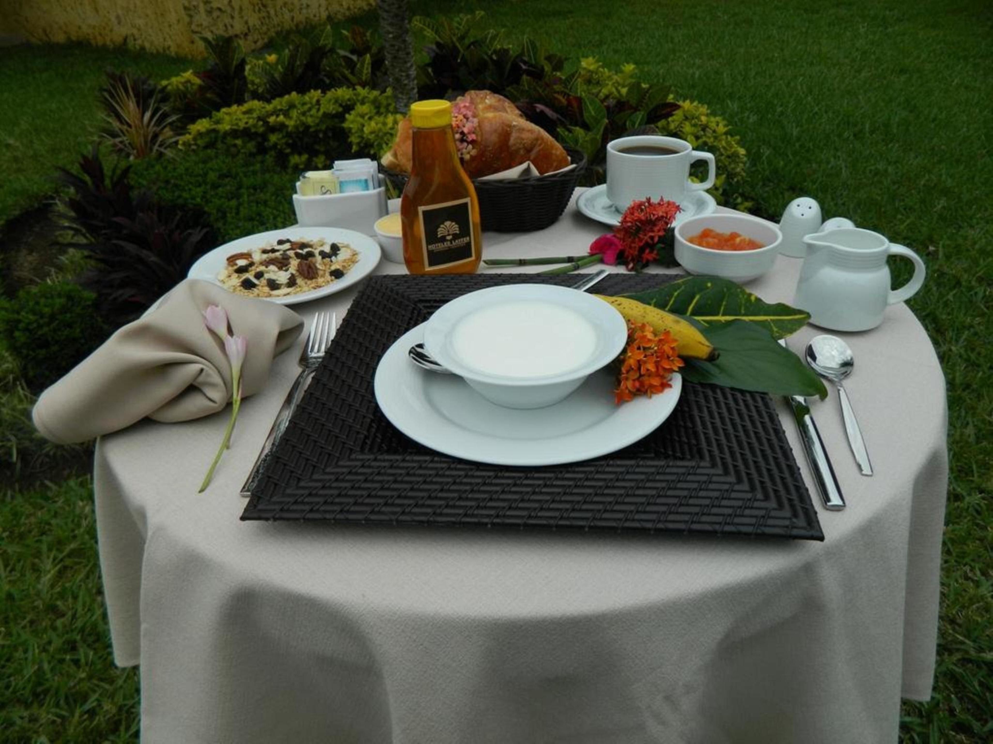 Une table dressée à l’extérieur, avec une assiette de nourriture, une tasse de café, une bouteille de miel et un bouquet de fleurs.