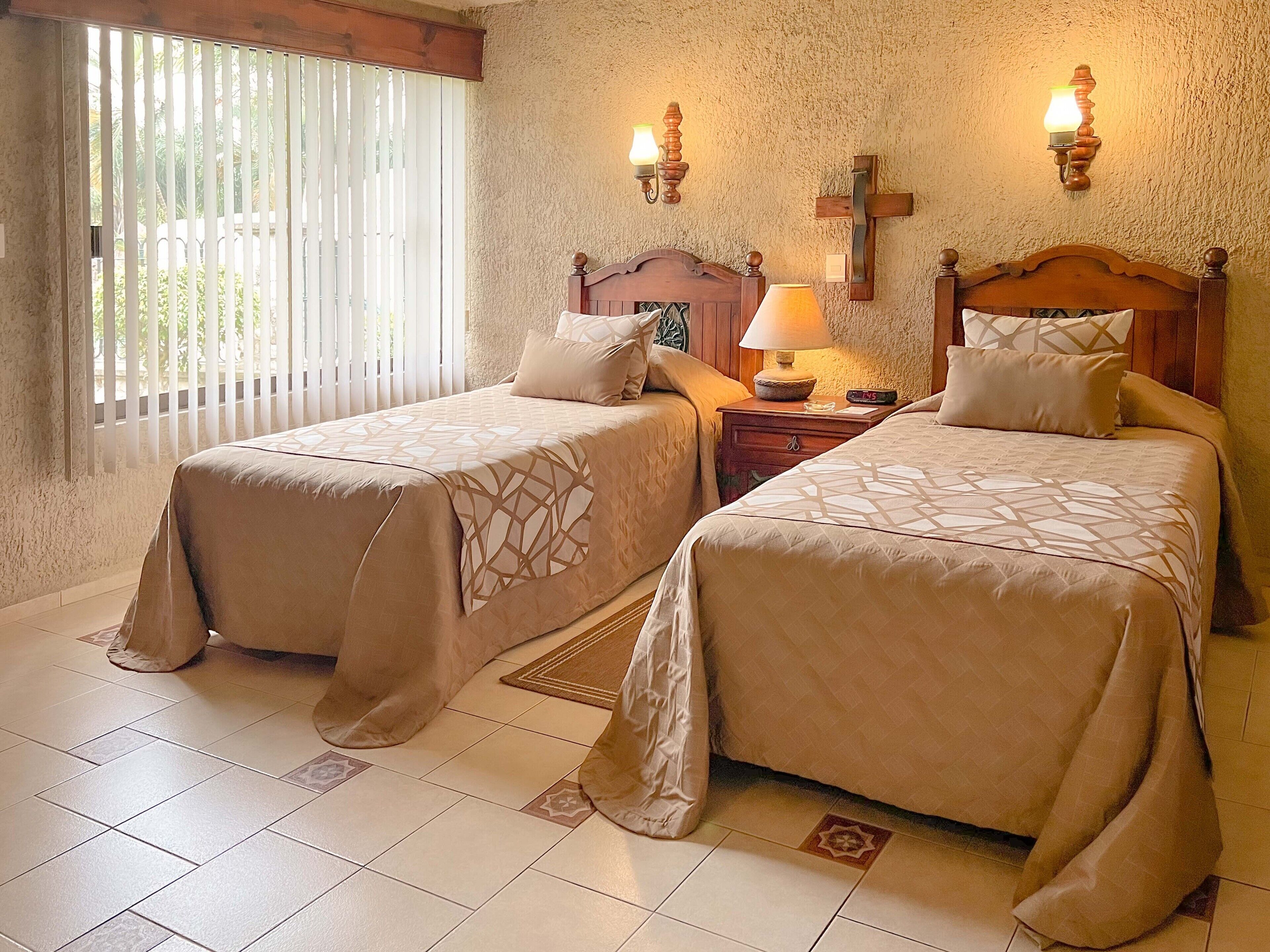 A hotel room with two beds, a wooden headboard, a nightstand with a lamp, and a window with blinds.