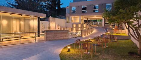 A modern building with a glass facade, outdoor seating area with chairs and tables, and a curved pathway leading to the entrance.