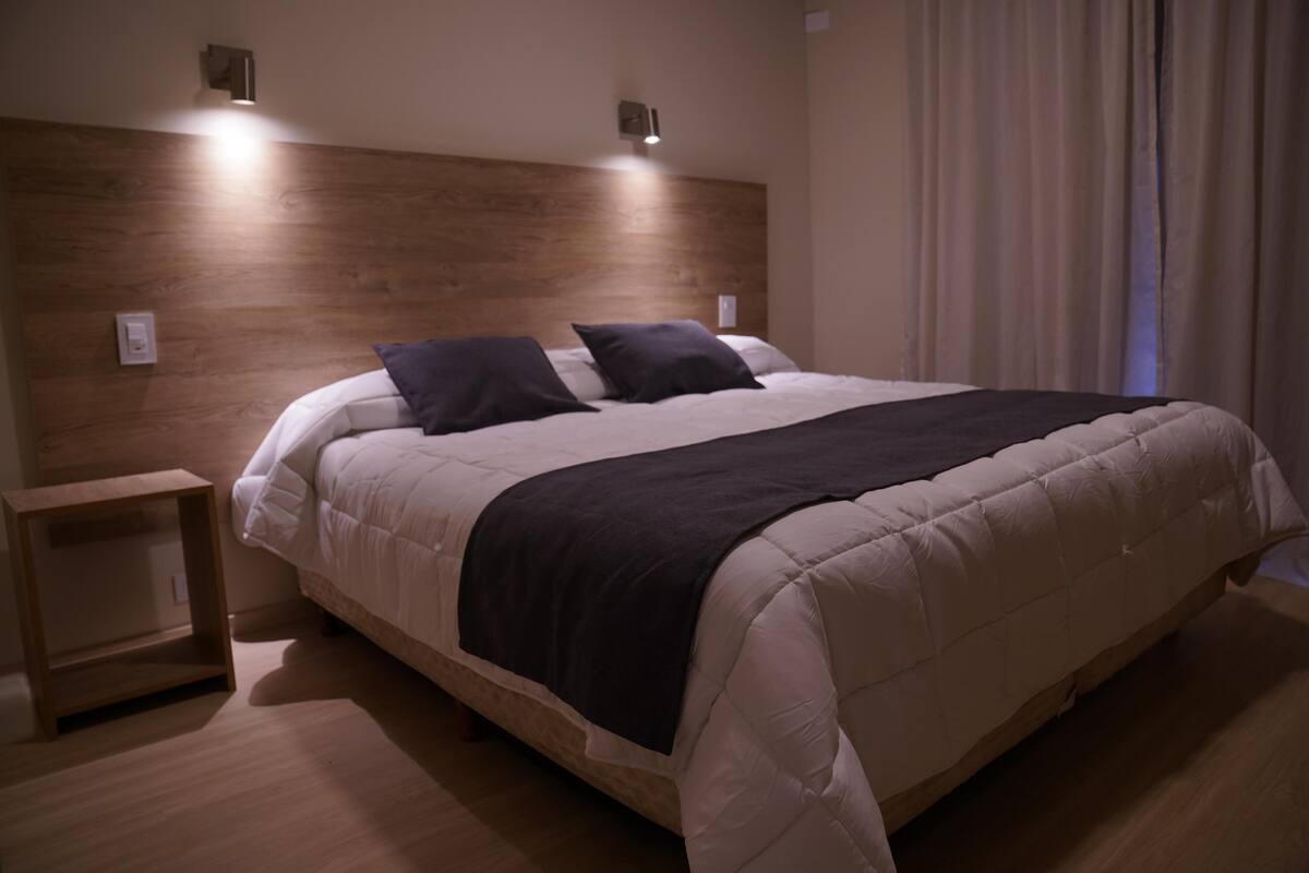 A neatly made bed with a dark blanket and two pillows, a wooden headboard, and bedside tables.