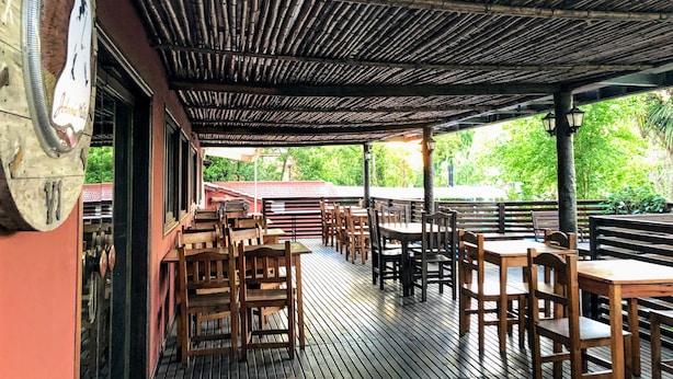 An outdoor patio with wooden tables and chairs, a bamboo ceiling, and a view of greenery in the background.