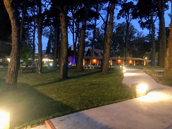 Una cabaña de diseño rústico, con un edificio central rodeado de árboles, un sendero pavimentado y zonas de estar al aire libre.