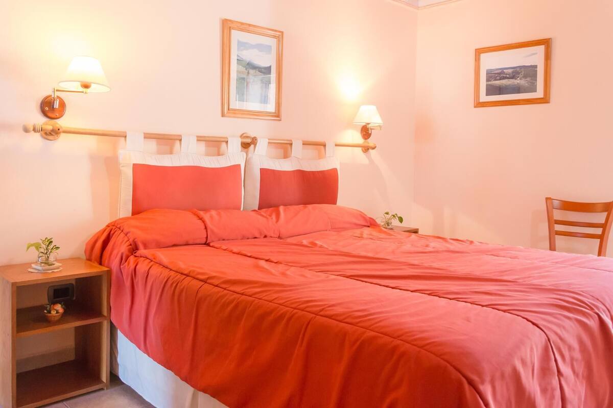 A neatly made bed with a red bedspread, two wall-mounted lamps, and two framed pictures on the wall.