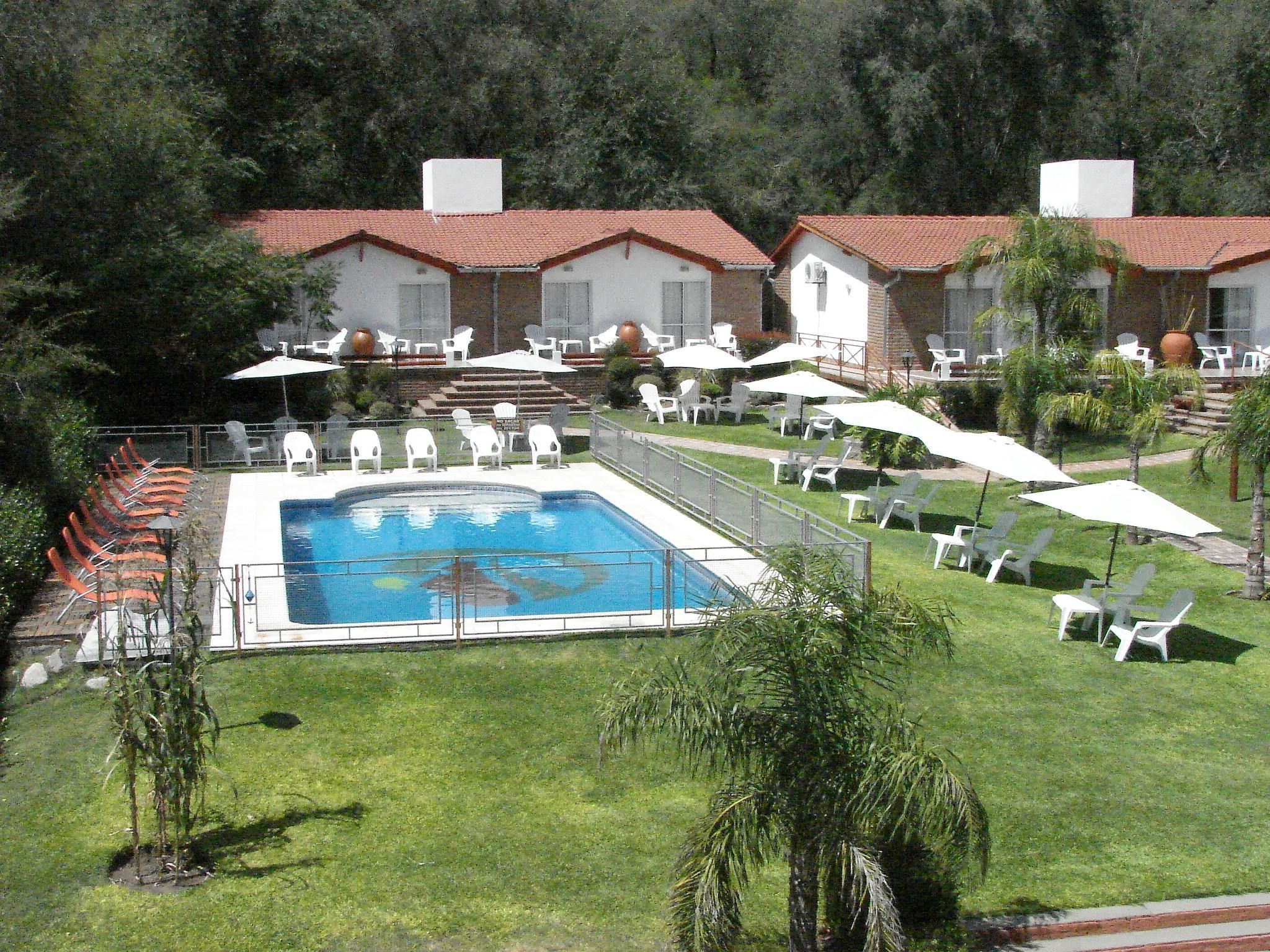 A pool area with a rectangular pool, surrounded by white lounge chairs and umbrellas, and a building with red-tiled roofs in the background.