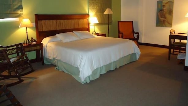 A hotel room with a large bed, two bedside tables with lamps, a wooden rocking chair, and a desk with a chair. - Hotel Casa del Sol (Managua)