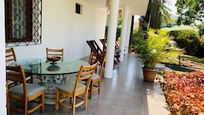 A covered patio with a glass table surrounded by wooden chairs, a potted plant, and a view of a garden with various plants and a small waterfall. - Hotel Casa del Sol (Managua)