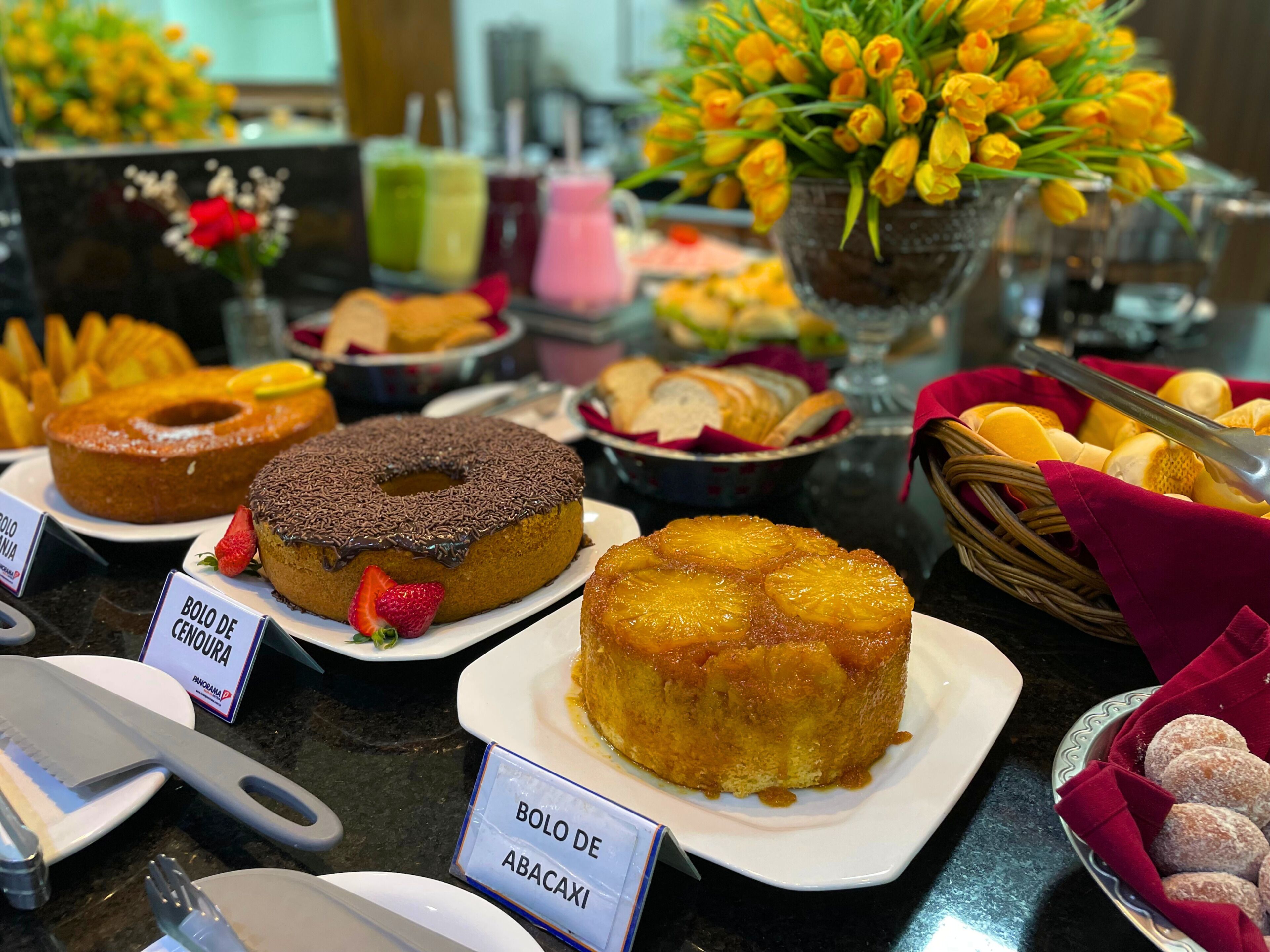 Um buffet com diversos doces, como um donut coberto de chocolate, um bolo invertido de abacaxi e um prato de biscoitos com açúcar de confeiteiro.