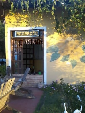 A reception area with a sign reading "RECEPCAO," a wooden bench outside, and a garden with flowers and plants. - Pousada dos Girassois (Prado)