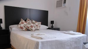 A neatly made bed with white linens and floral pillows, a bedside table with a lamp, and a chair with a black and white cushion. - Ñanderoga (Corrientes)