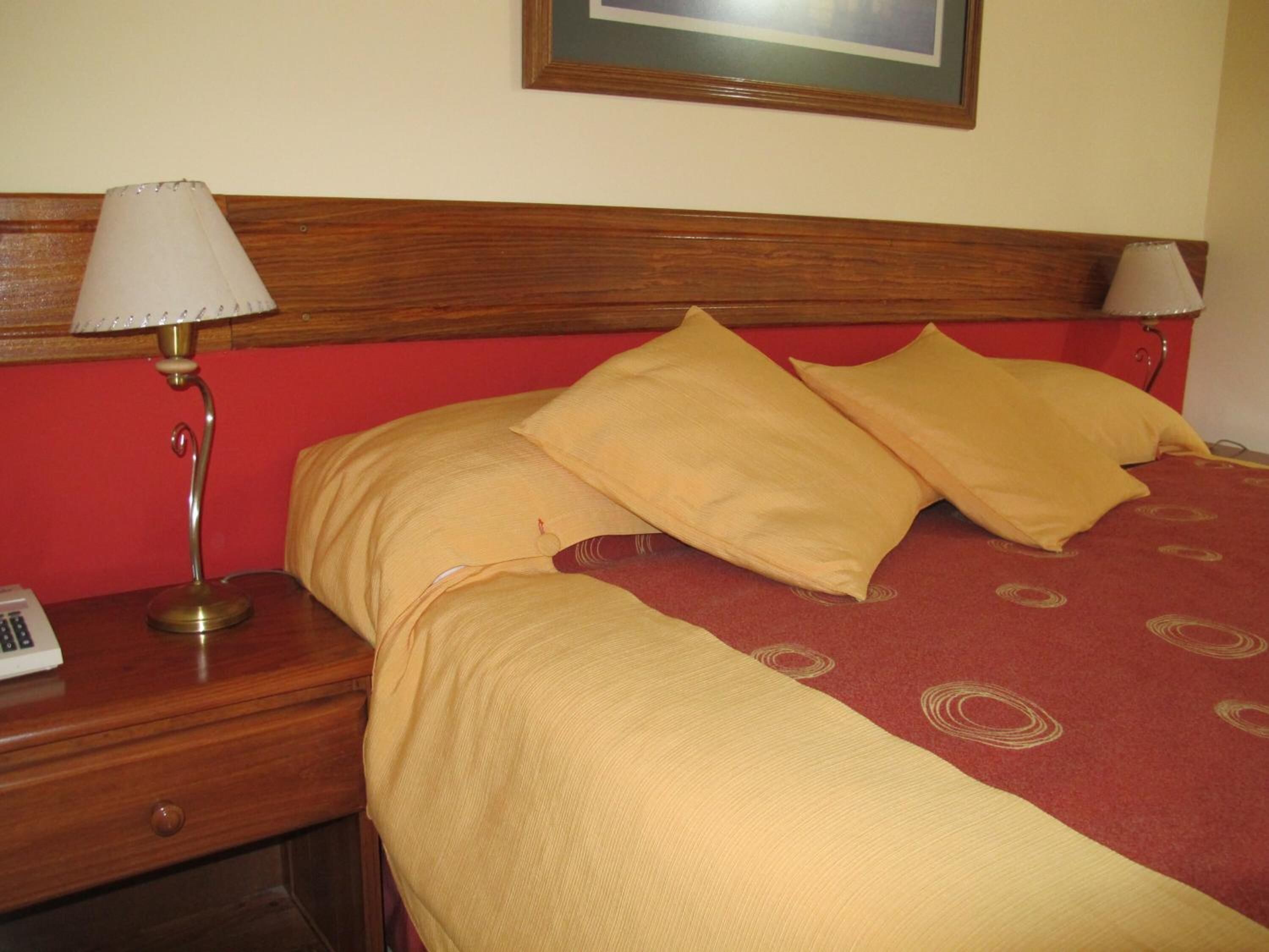 Habitación de hotel con cabecera de madera, cama con ropa de cama amarilla y mesita de noche con lámpara y teléfono.