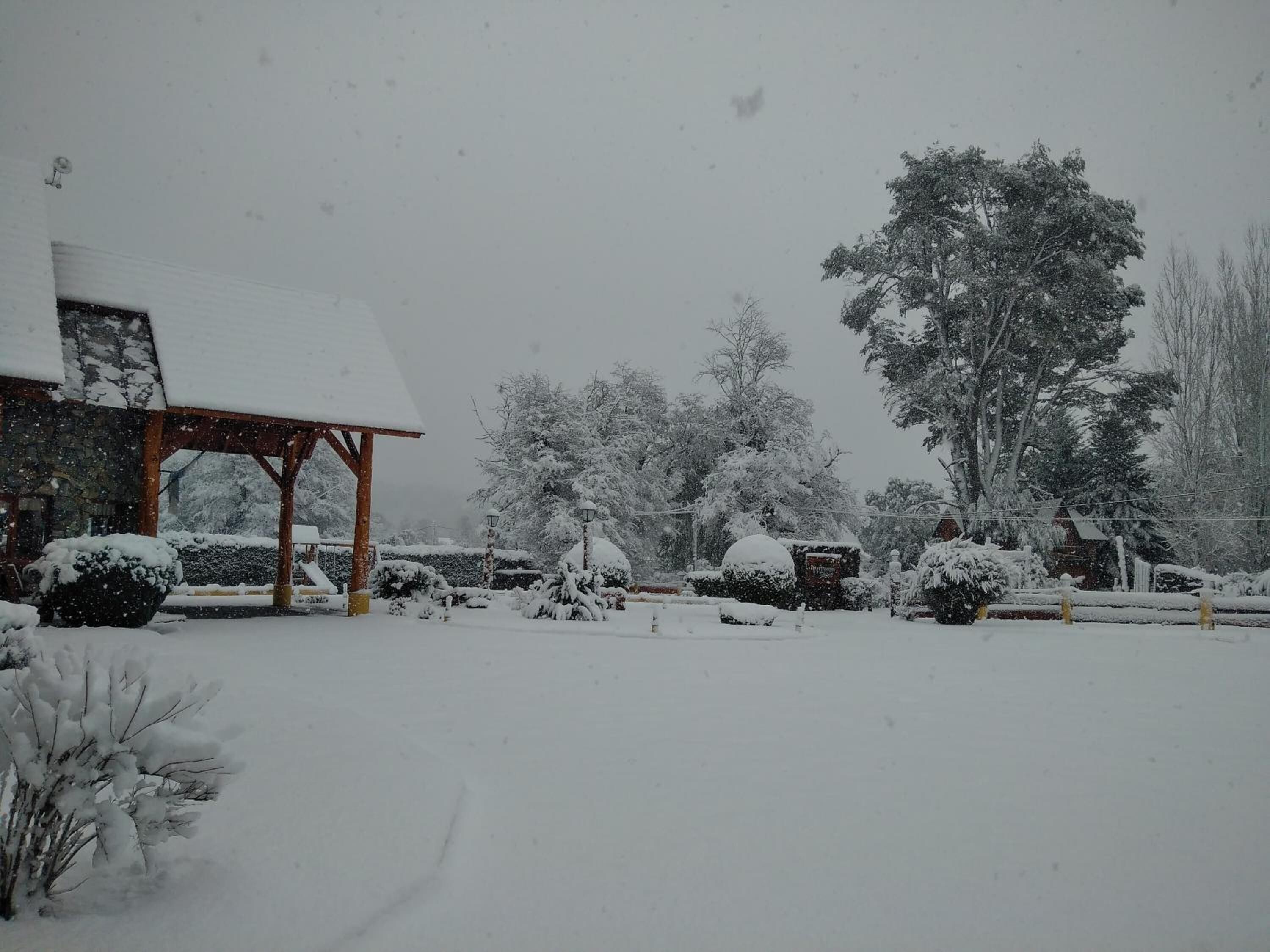 Un espacio exterior cubierto de nieve con un pabellón de madera, árboles nevados y un edificio de piedra.