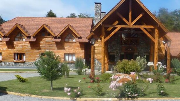 Un bâtiment de style chalet en rondins de bois, doté de plusieurs pièces, d’une cheminée en pierre et d’un jardin aménagé avec des fleurs et des arbres.