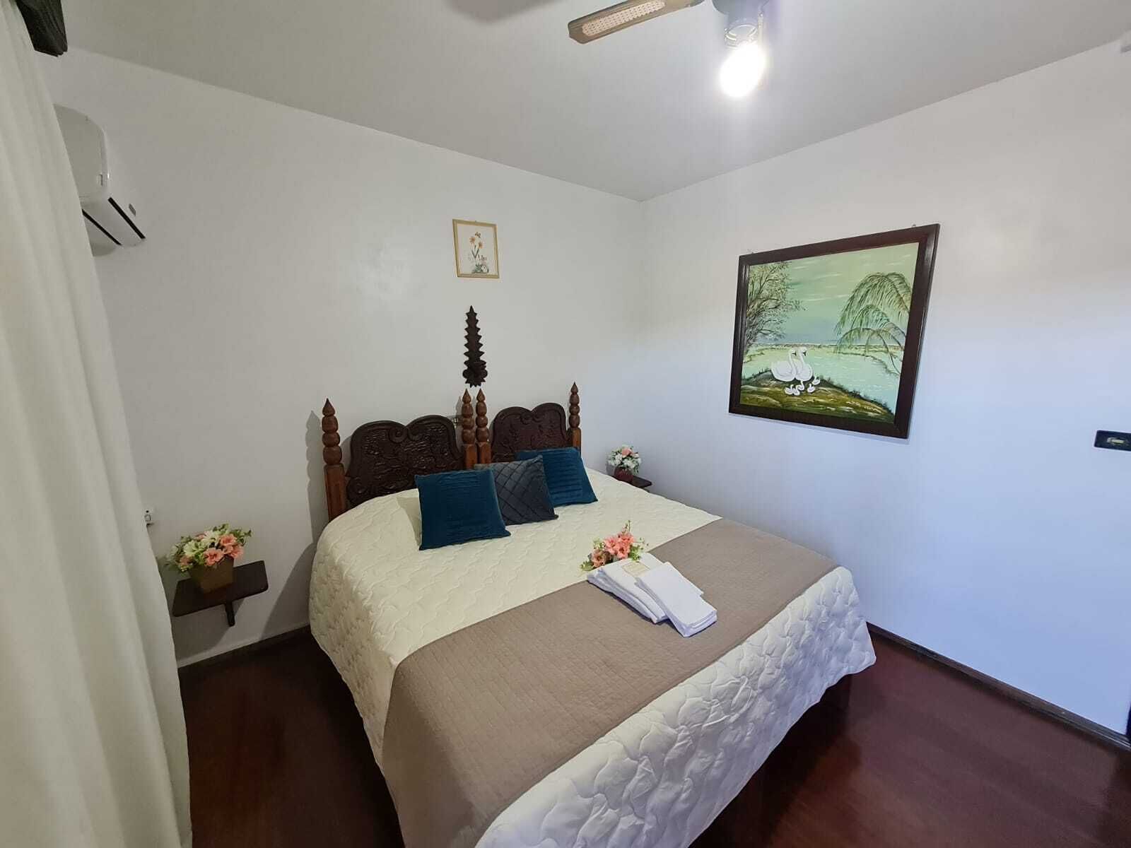 Una cama bien hecha con cabecera de madera, un cuadro de paisaje enmarcado en la pared y una mesita de noche con un florero.