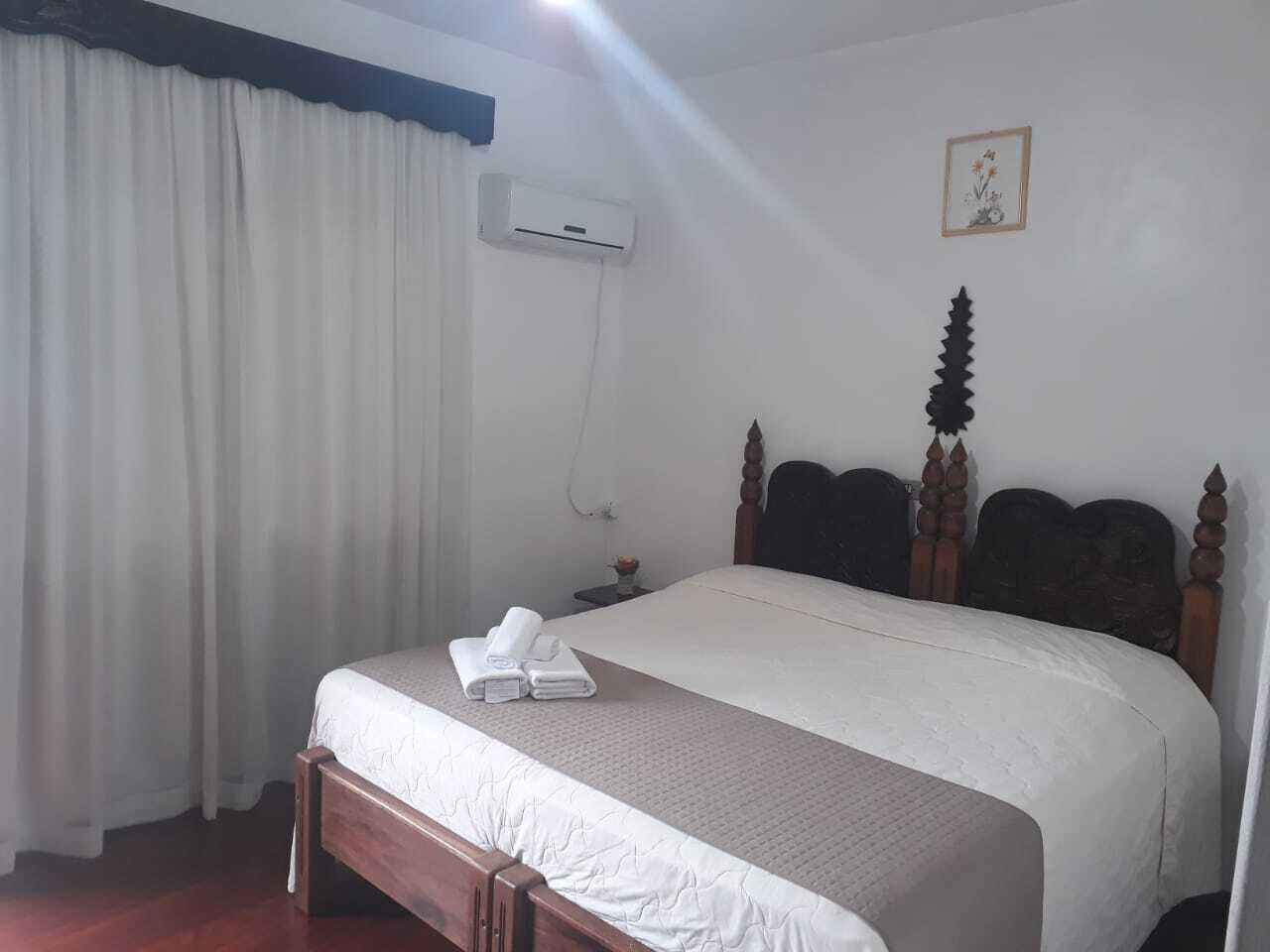 A hotel room with a wooden bed, white curtains, an air conditioning unit, and a small bedside table with a vase of flowers.