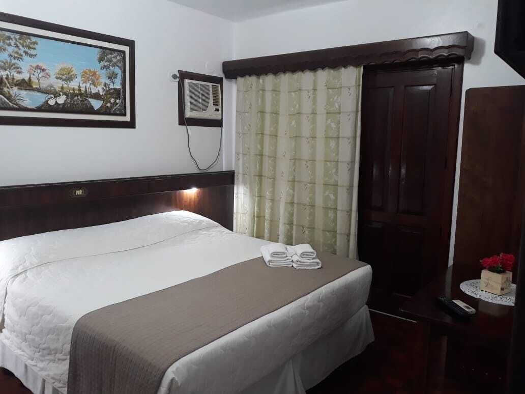 Una habitación de hotel con una cama grande, un cuadro en la pared, un equipo de aire acondicionado, una cortina, una puerta de madera y una pequeña mesa con un jarrón de flores.