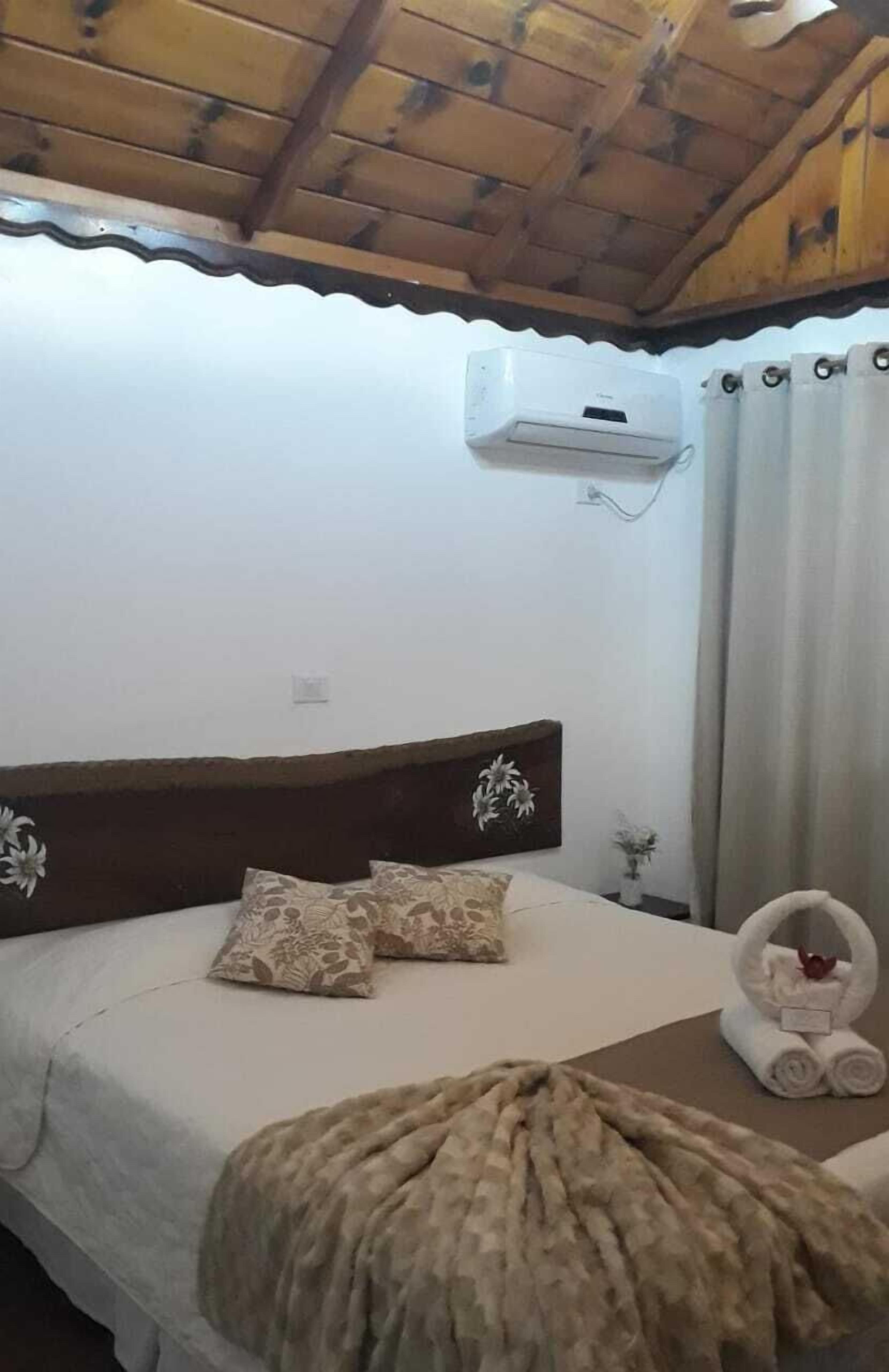 Un dormitorio con techo de madera, una cama con almohadas estampadas, un aire acondicionado de pared y una mesita con toallas.