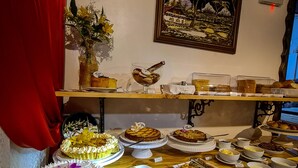 Una mesa de buffet con diversos platos y postres, un cuadro enmarcado de un paisaje, y una cortina roja.