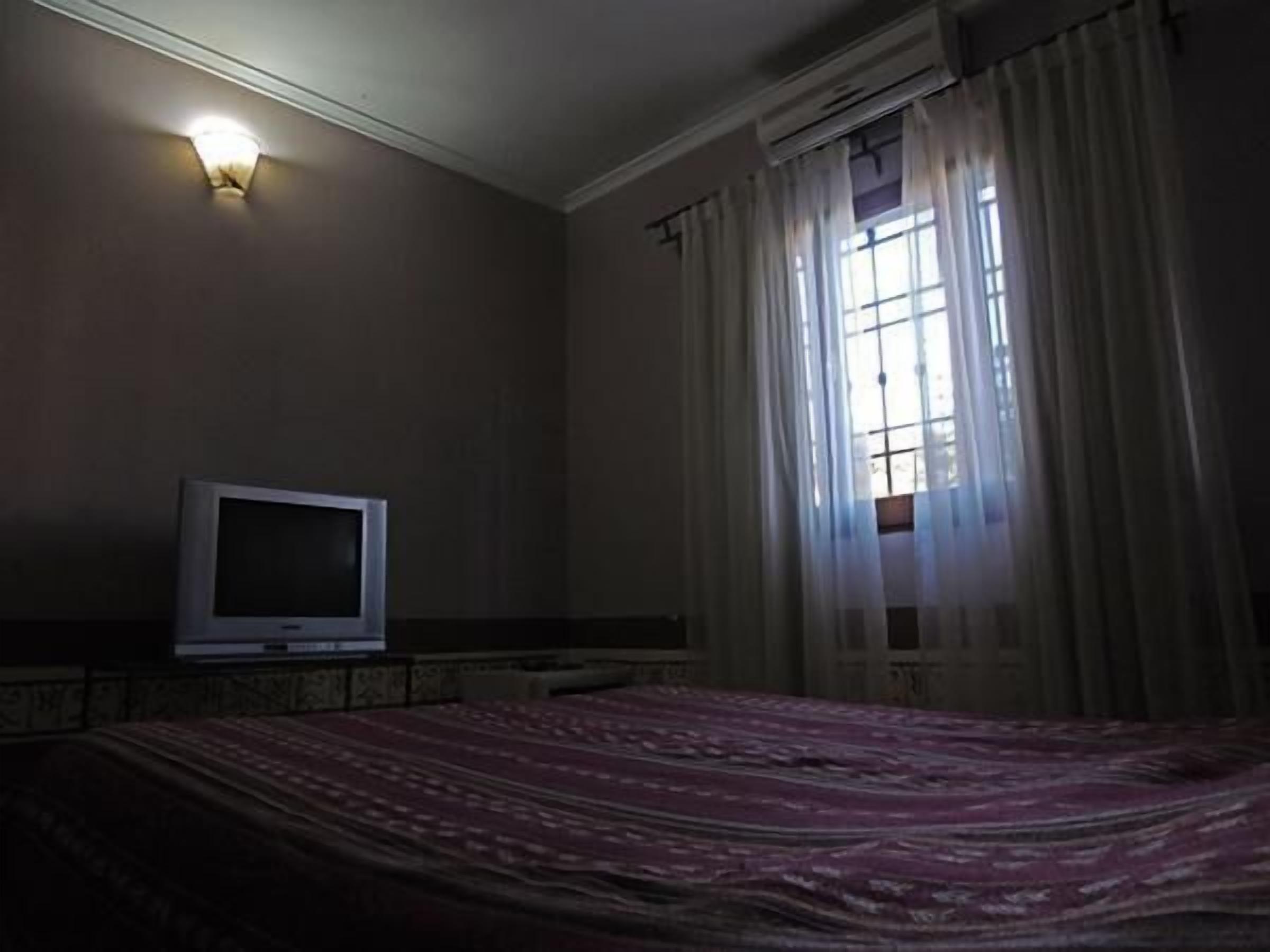 Una habitación con poca luz, una cama, un televisor y un ventanal con cortinas.