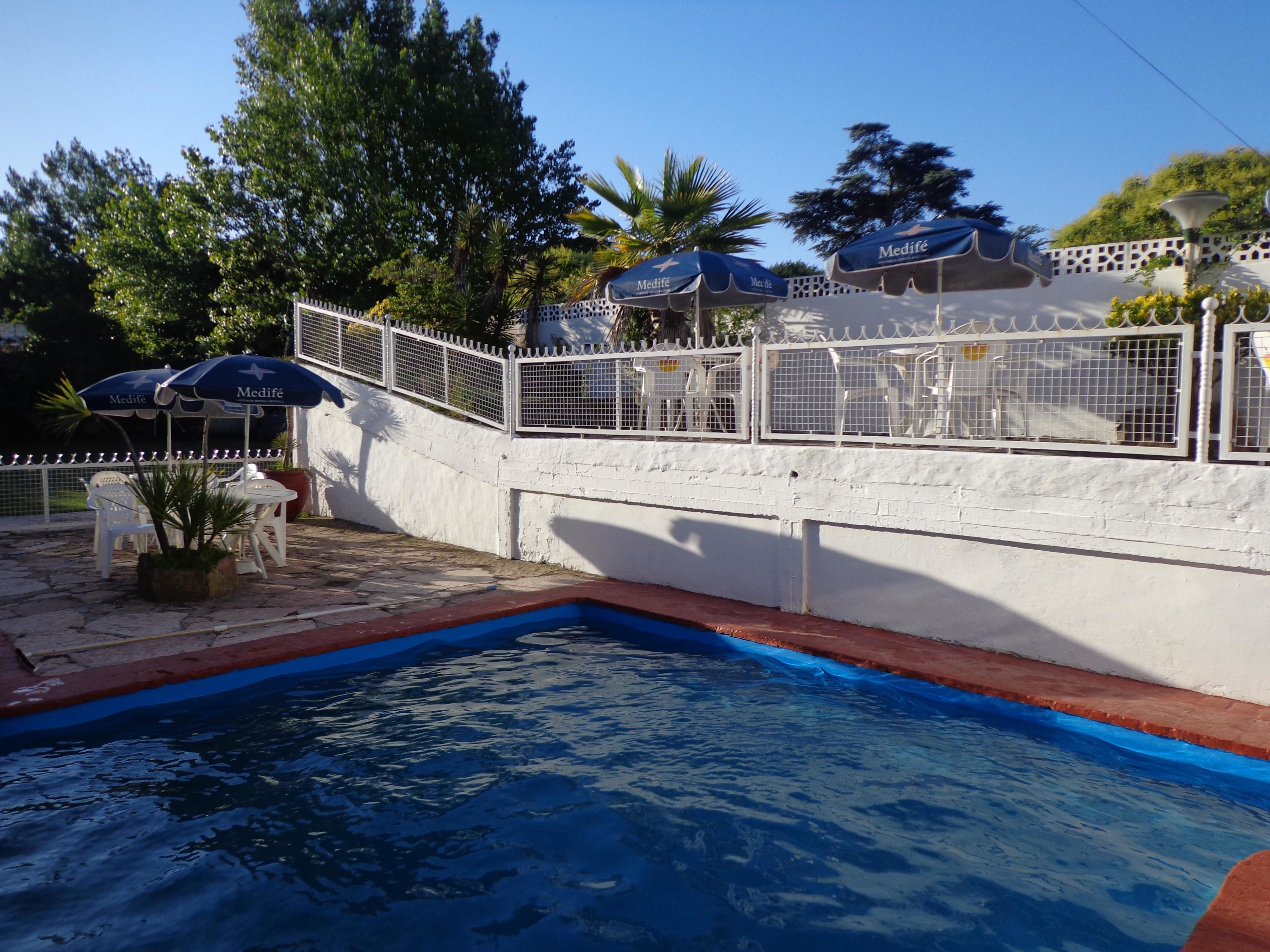 Una piscina con pared blanca y detalles azules, rodeada de mobiliario exterior y sombrillas.
