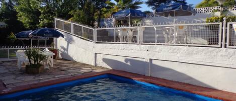Una piscina con pared blanca y detalles azules, rodeada de mobiliario exterior y sombrillas.