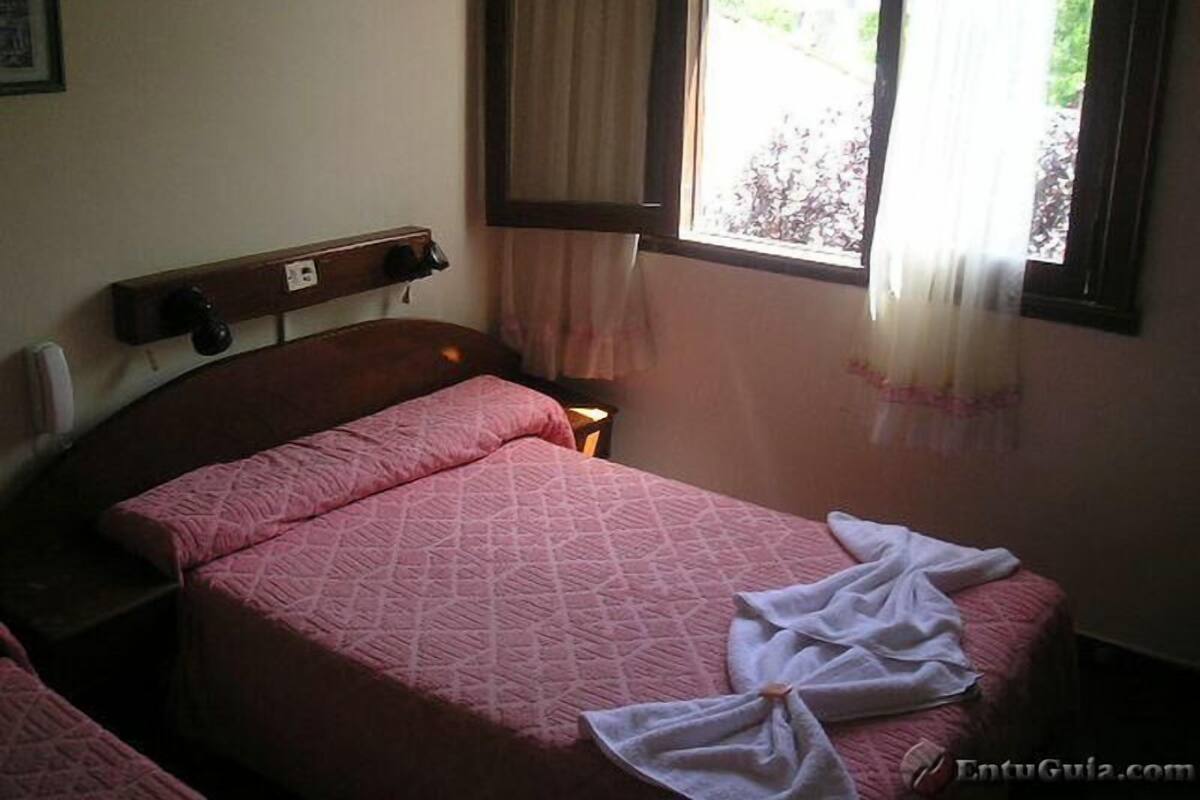 Un lit simple avec une couverture rose, une table de chevet, une fenêtre avec des rideaux et un téléphone fixé au mur.