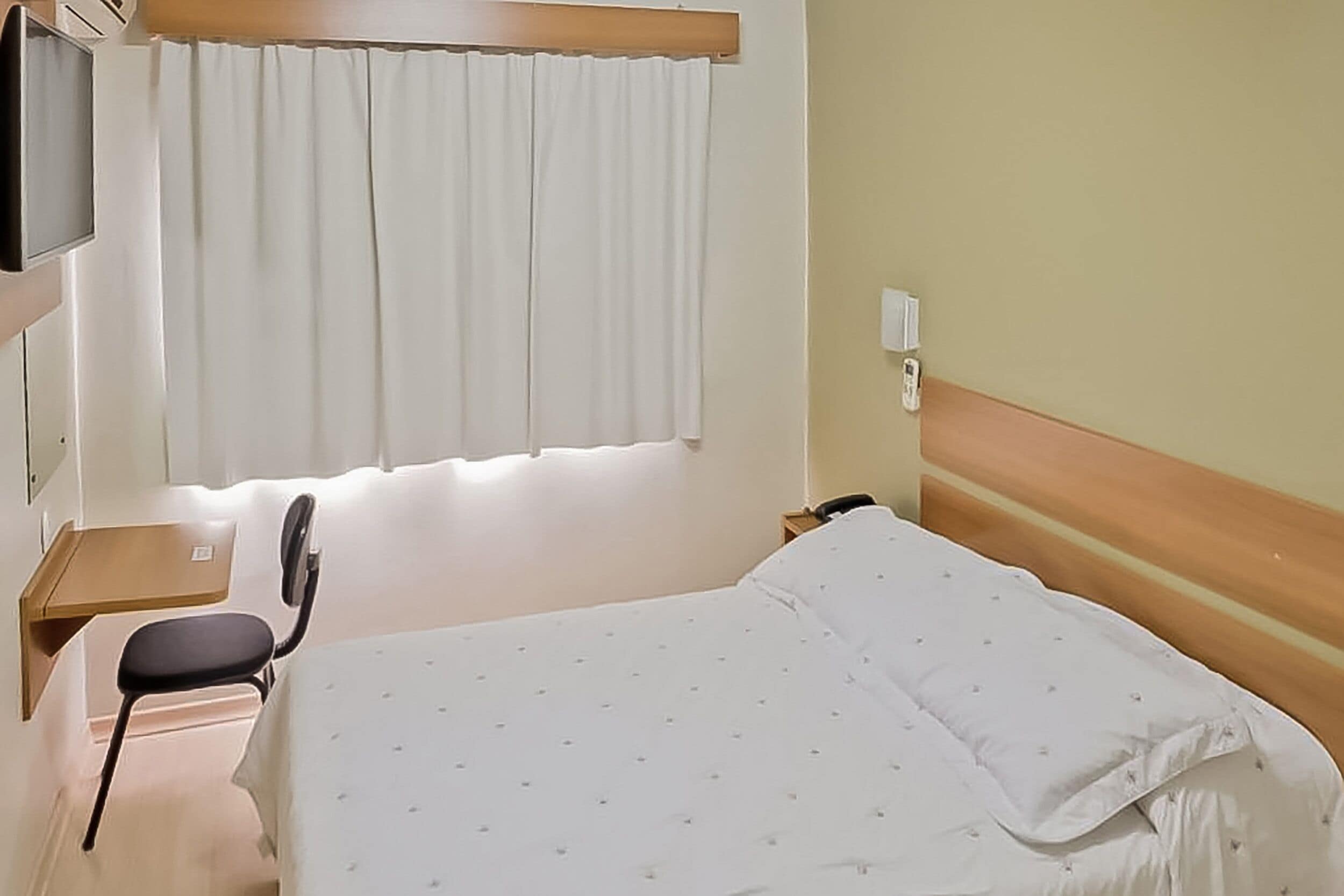 Habitación de hotel con cama, escritorio, silla, televisión y ventana con cortinas.