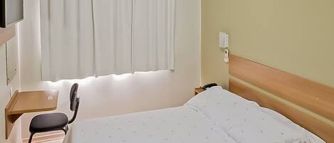 Habitación de hotel con cama, escritorio, silla, televisión y ventana con cortinas.