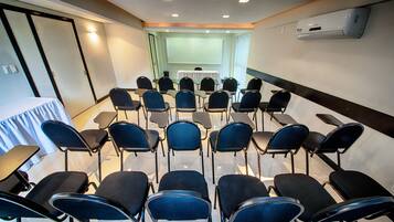 Un salón de conferencias con filas de sillas, una pantalla para proyector y una mesa cubierta con un mantel blanco.